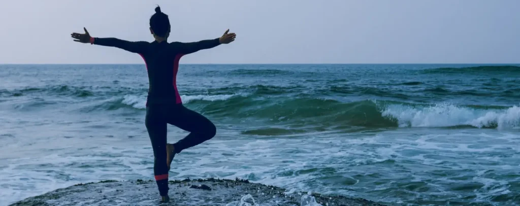 Person in Yoga-Balancepose auf einem Felsen am Meer, mit ausgebreiteten Armen und Blick auf die Ostsee, sanfte Wellen im Vordergrund.
