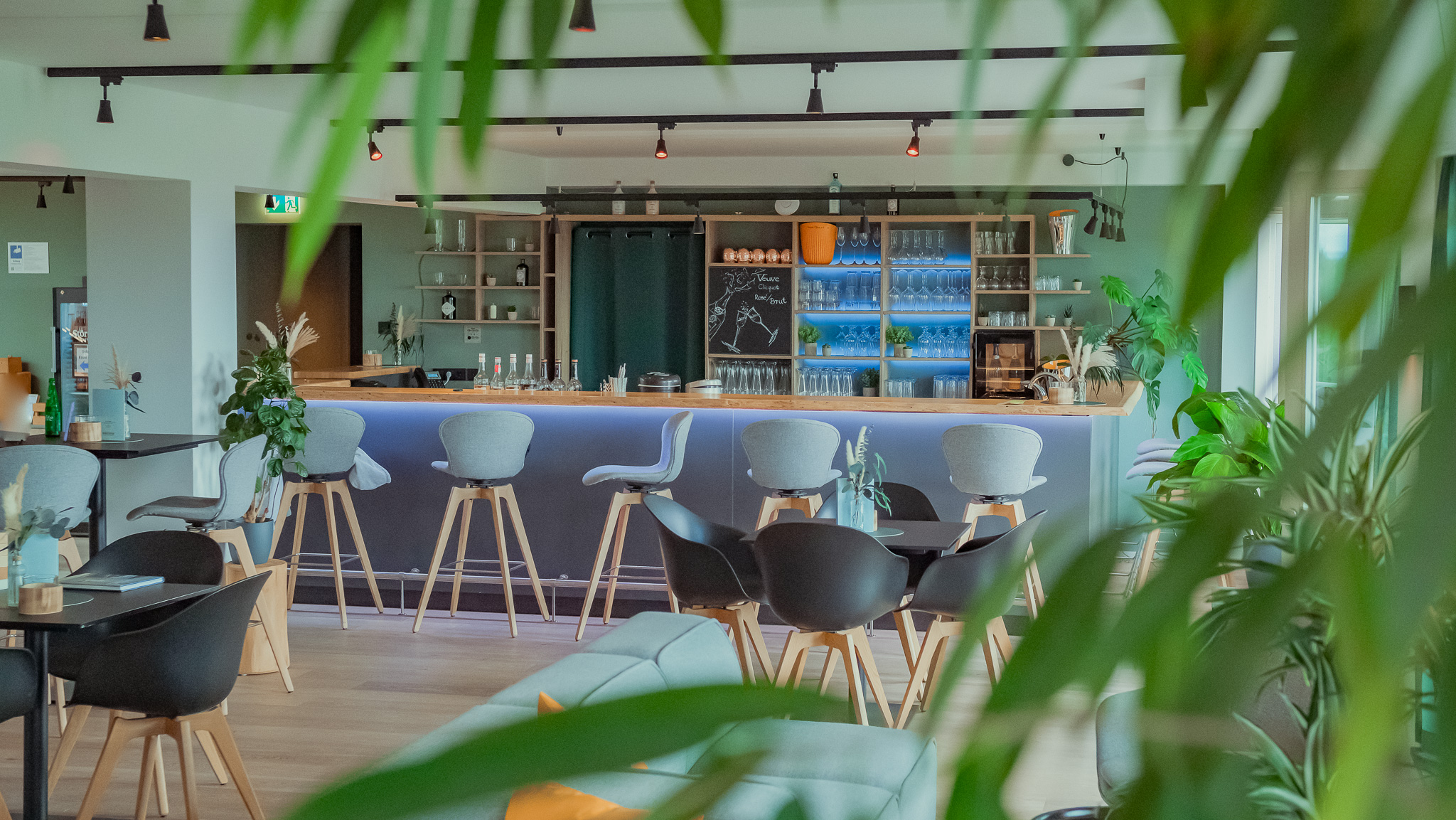 Skybar in der Bergstation Prora - Mariandl am Meer Ein Bartresen einer Bar mit grauen Barhockern, Pflanzenelementen und einem Tisch mit vier schwarzen Stühlen.