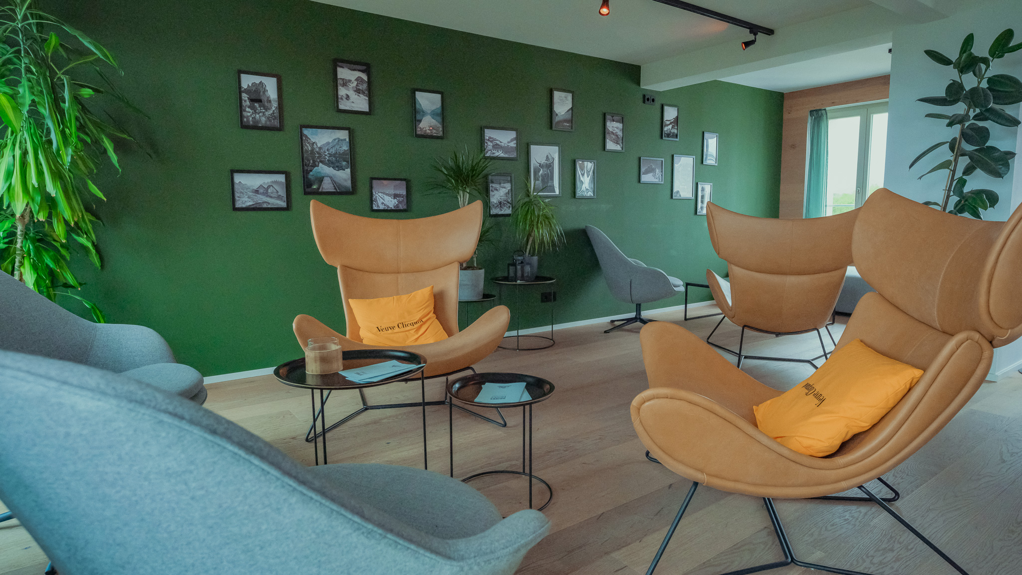Bergstation Fotowand - Mariandl am Meer Ein Ambiente in einer Bar, mit einer grünen Wand, auf der verschiedene Bilderrahmen hängen. Drei Disign-Sessel und zwei Beistelltische stehen im Raum.