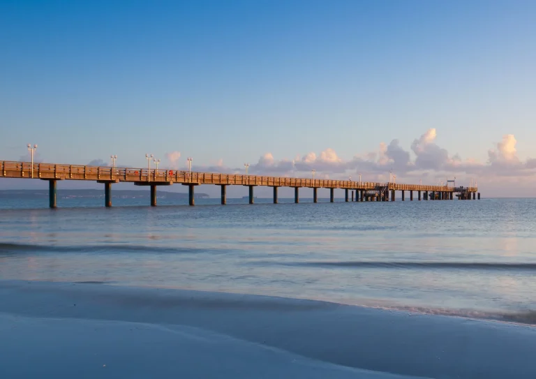 Seebrücke ragt vom Sandstrand weit hinaus in die ruhige Ostsee, beleuchtet vom warmen Licht der untergehenden Sonne unter klarem Himmel.